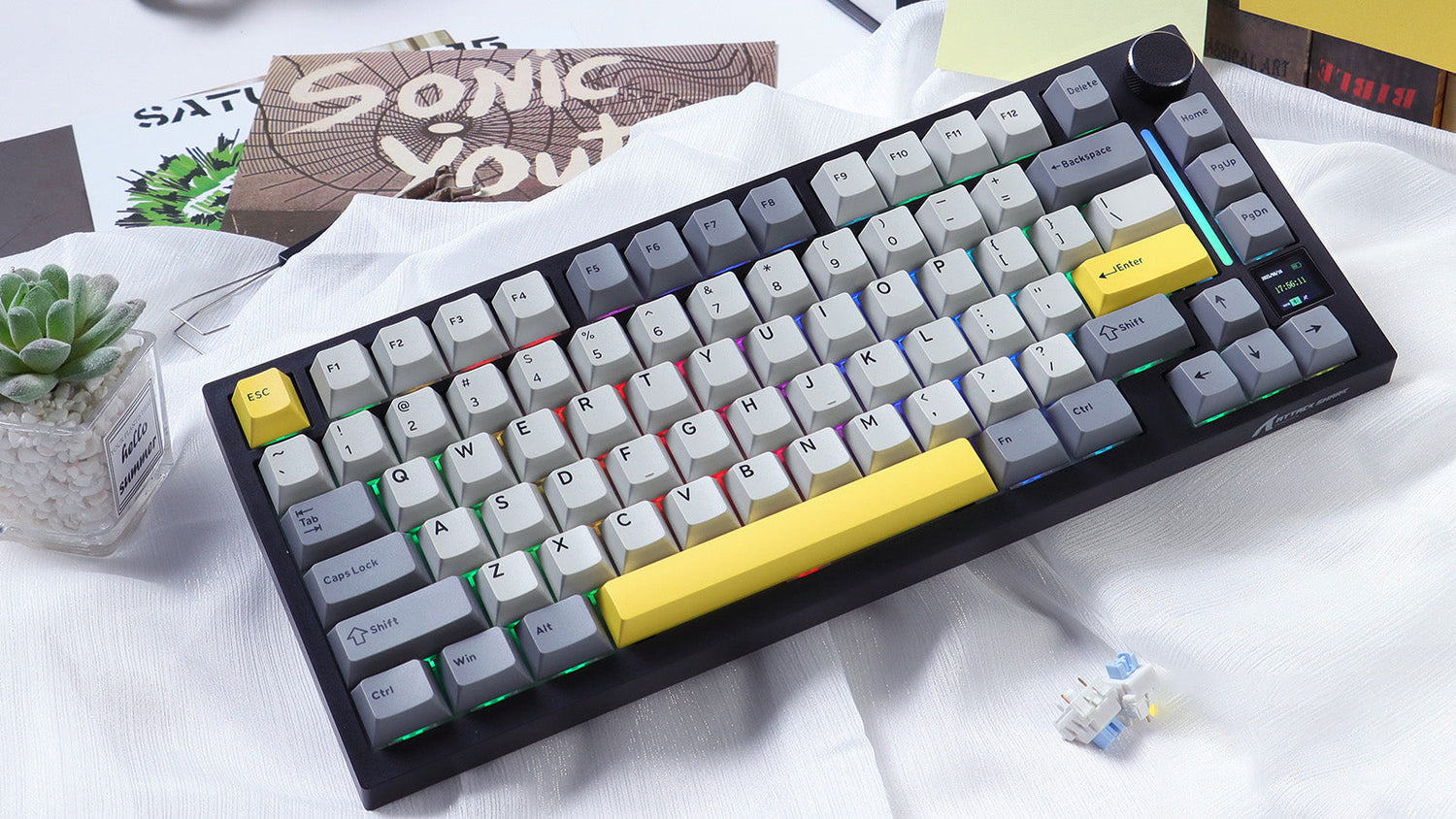 Compact mechanical keyboard with grey and yellow accent keycaps on a white desk, featuring RGB lighting and positioned next to a small succulent plant and a 'Sonic Youth' album cover